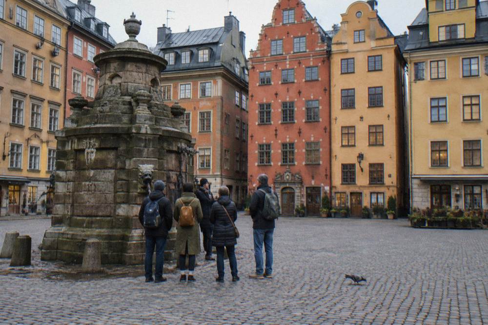 A guide speaking to a small group of visitors in a Stockholm Old Town square lined with colorful old buildings