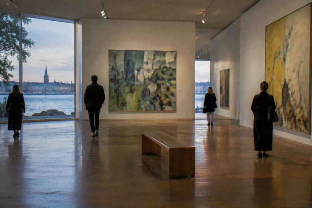 A museum gallery interior lit for evening visiting, with paintings on the walls and a few visitors walking through