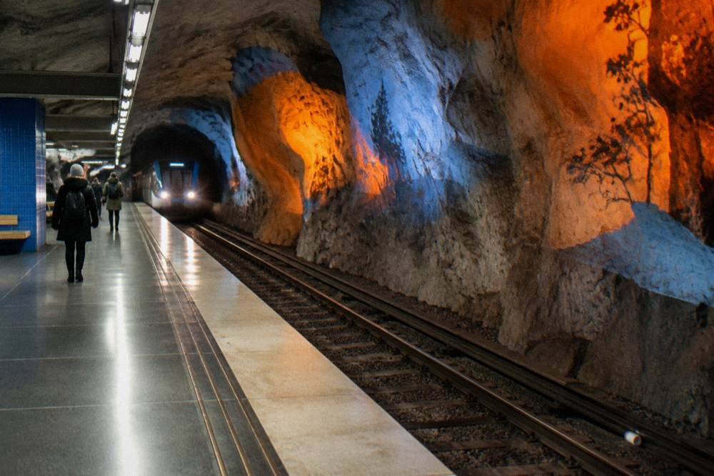 A blue-line Stockholm metro platform with exposed rock walls and colorful artwork along the tunnel