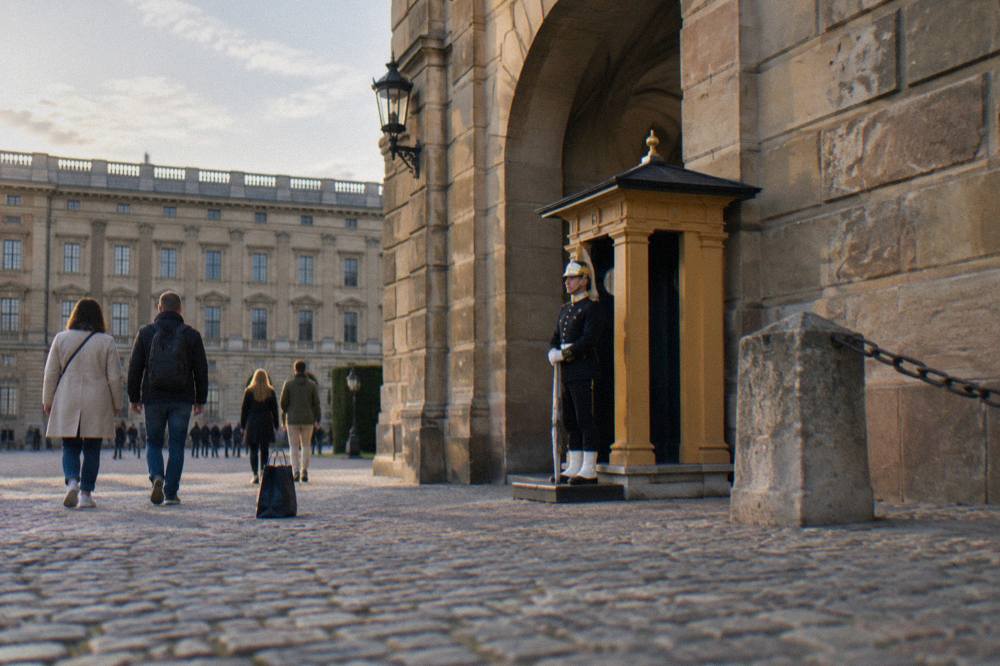 A royal guard standing at a post beside an arched gateway of the Stockholm Royal Palace, with visitors walking past in the courtyard