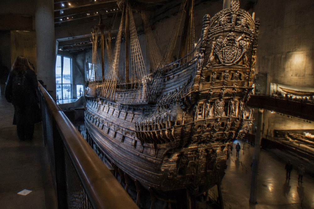 The tall dark wooden hull of the Vasa warship inside the museum hall, seen from a walkway