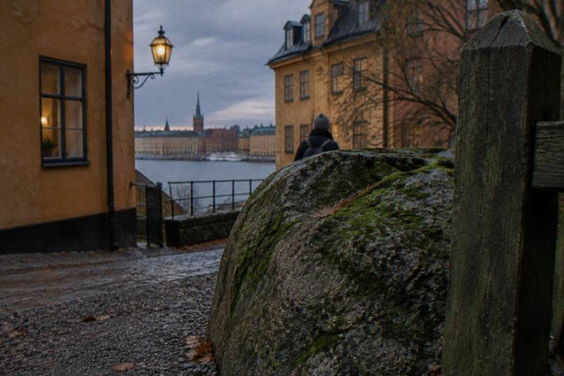 Ancient carved runestone standing beside a coastal path near Stockholm, Sweden, pale granite etched with Viking age inscriptions, pine trees behind, calm grey water and a guided group pausing to listen.