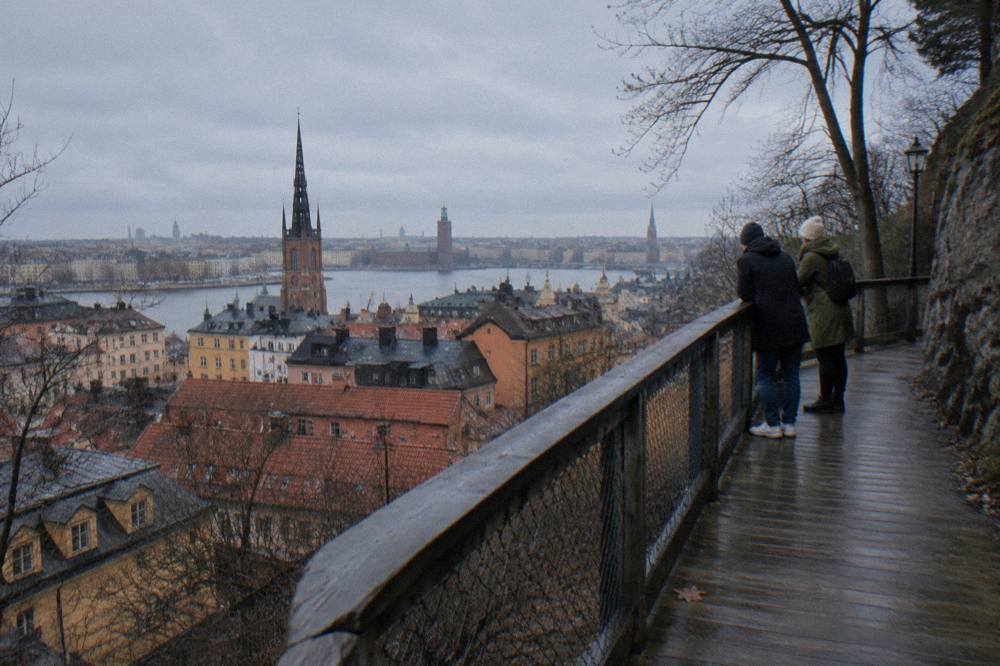 People walking a narrow cliffside path above red rooftops and a church spire in Stockholm