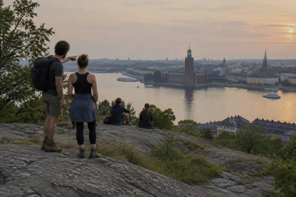 Walkers pausing on a wooden cliffside boardwalk above a city skyline and water